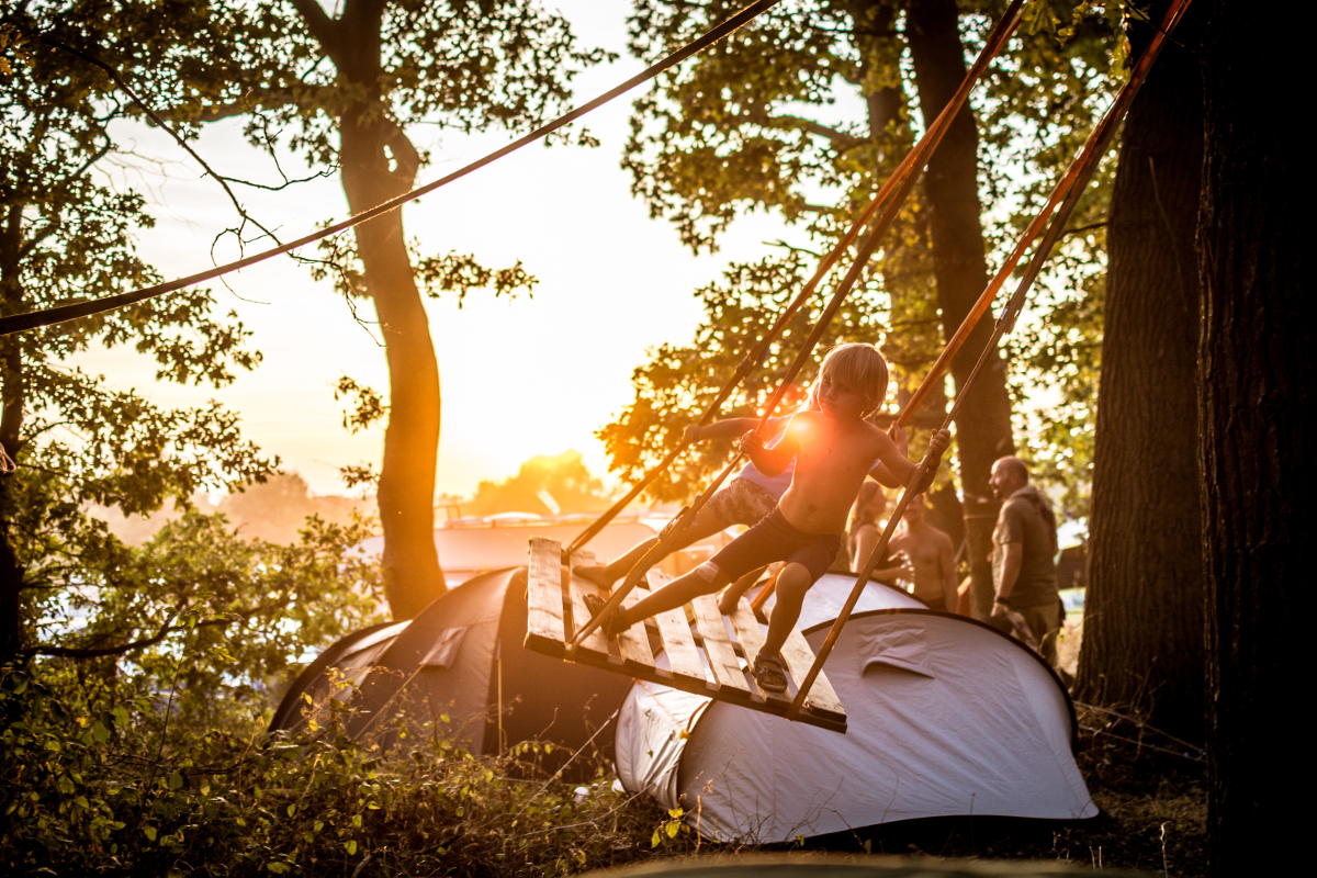 Im Hintergrund ist ein Sonnenuntergang und ein paar Zelte. Zwei Kinder schaukeln im Stehen.