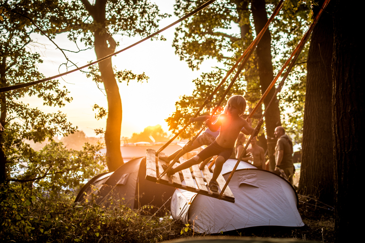 Camping Im Hintergrund ist ein Sonnenuntergang und ein paar Zelte. Zwei Kinder schaukeln im Stehen.