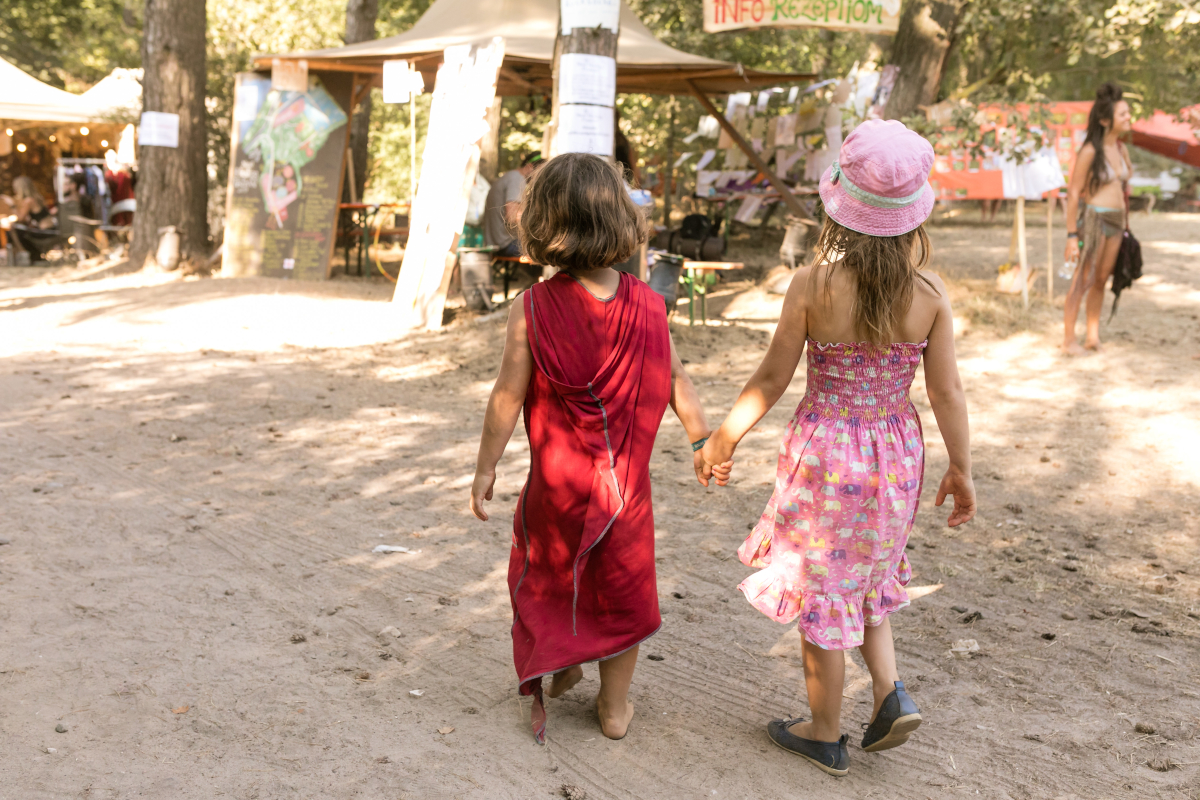 Eltern-Kinder Zwei Kinder von hinten fotografiert, die Hand in Hand über das New Healing festival laufen. Das linke Kind trägt ein rotes Kleid. Das rechte kind trägt ein rosafarbenes Kleid und einen rosa Hut.