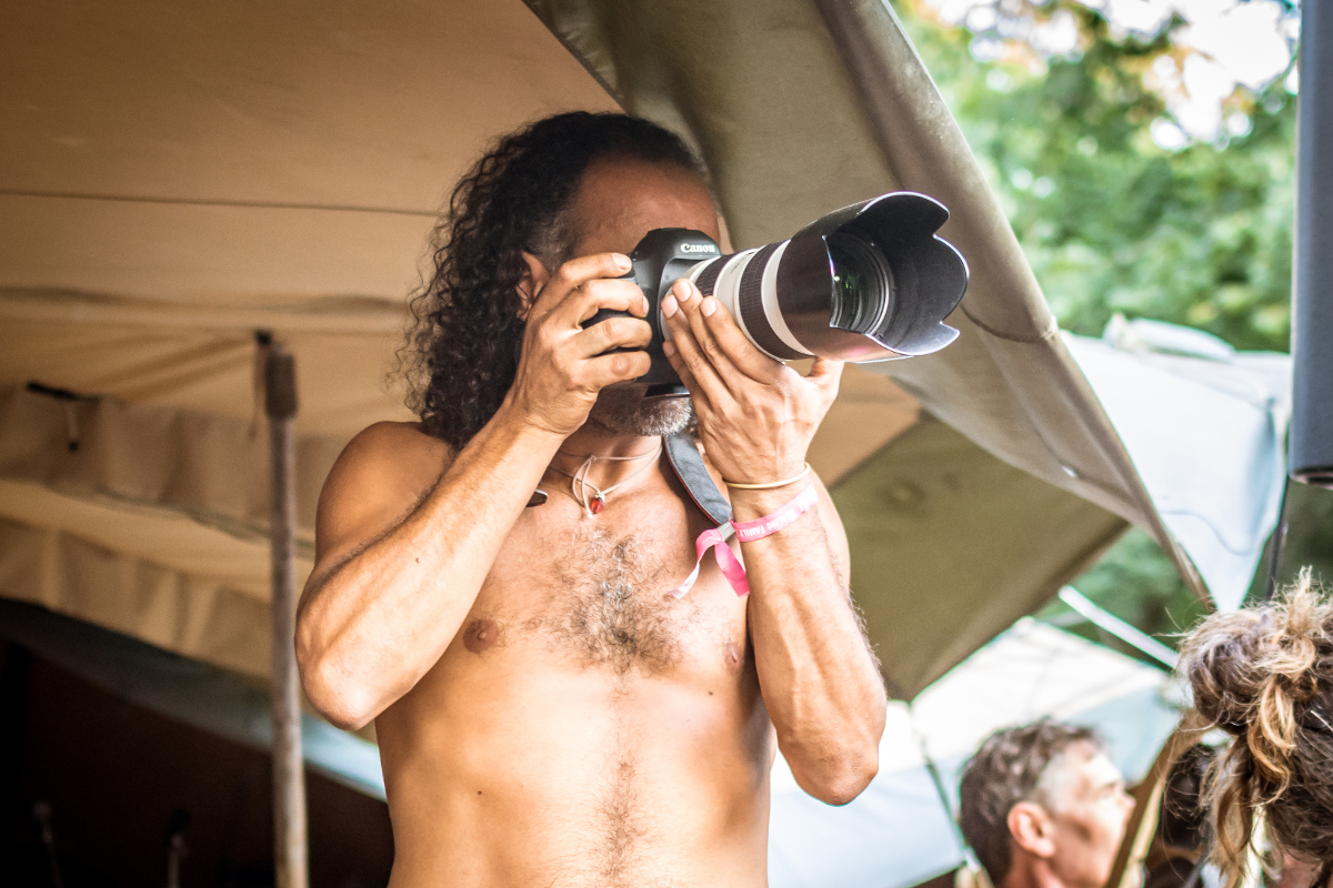 Fotografieren Ein Mann mit mittellangen, gelockten Haaren hält seine Kamera vorm Gesicht, um etwas zu fotografieren.