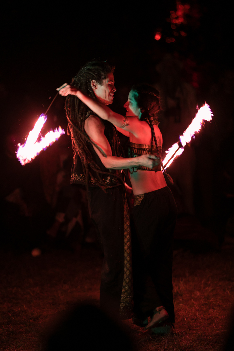 equilibra-feuer Ein Mann und eine Frau stehen sich mit den Gesichtern zugewandt gegenüber. Sie halten hinter dem Rücken des jeweils anderen eine brennende Schnur. Es ist Nacht