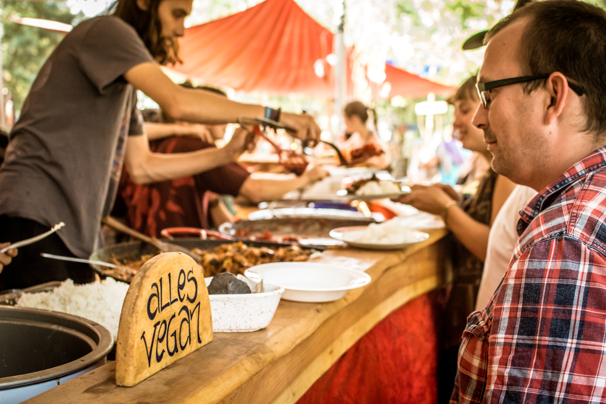 freiraum-der-einheit-ausgabe Eine Theke hinter der 3 Menschen Essen ausgeben. Auf der Theke sind ein paar Teller platziert und ein Holzschild auf dem alles vegan steht.
